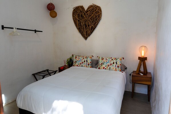 A bed with a white bedspread, two colorful pillows, a heart-shaped wall decoration, a bedside lamp, and a small table with a vase and a red cloth.