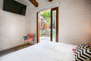 Une chambre avec un mur en briques blanches, un lit avec un matelas blanc, une chaise rouge et une vue sur un espace extérieur coloré avec des escaliers.