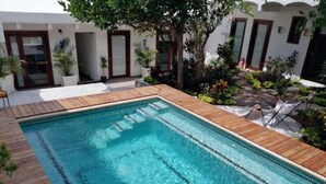 Une piscine rectangulaire entourée d’une terrasse en bois, située devant un bâtiment blanc doté de nombreuses portes et fenêtres.