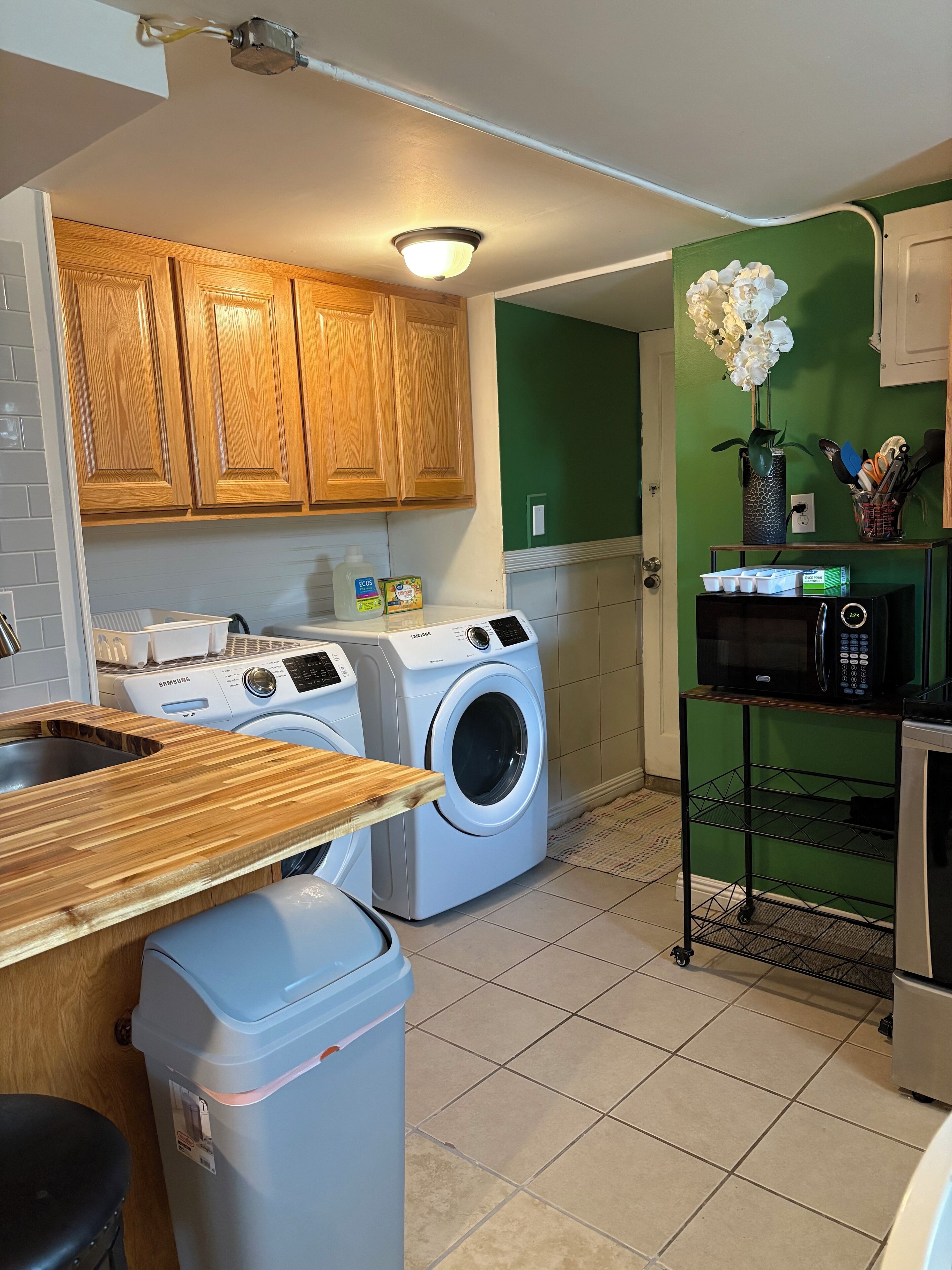 A compact kitchen with wooden cabinets, a washing machine, a microwave, and a trash bin.