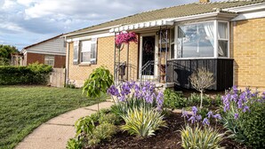 Une maison à deux étages, construite en briques, avec un porche à l’avant, un petit jardin parsemé de fleurs violettes et un ciel dégagé.