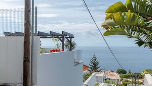 A coastal residential area with a white building, a view of the sea, and a balcony with red flowers. - GuestReady - Anakaj House (Funchal)