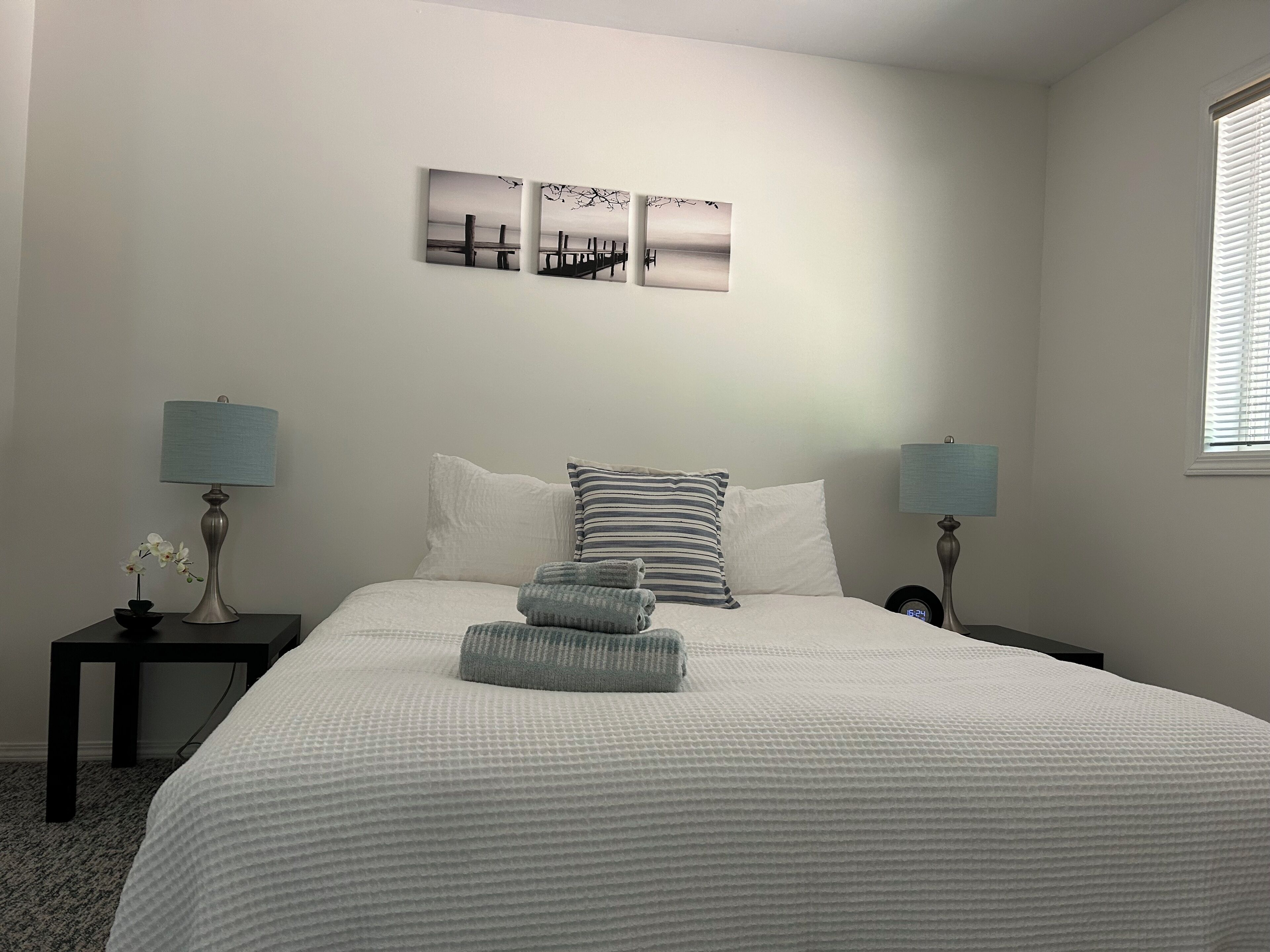 Un dormitorio con una cama, dos mesitas de noche, un ventanal y tres cuadros en la pared.