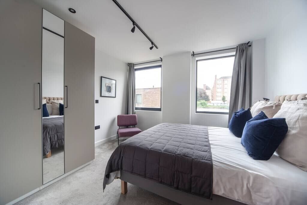 Un dormitorio con cama, una silla, armario y dos ventanas.