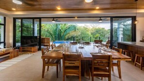 A spacious dining area with a long wooden table set for a meal, surrounded by large windows offering a view of lush greenery.