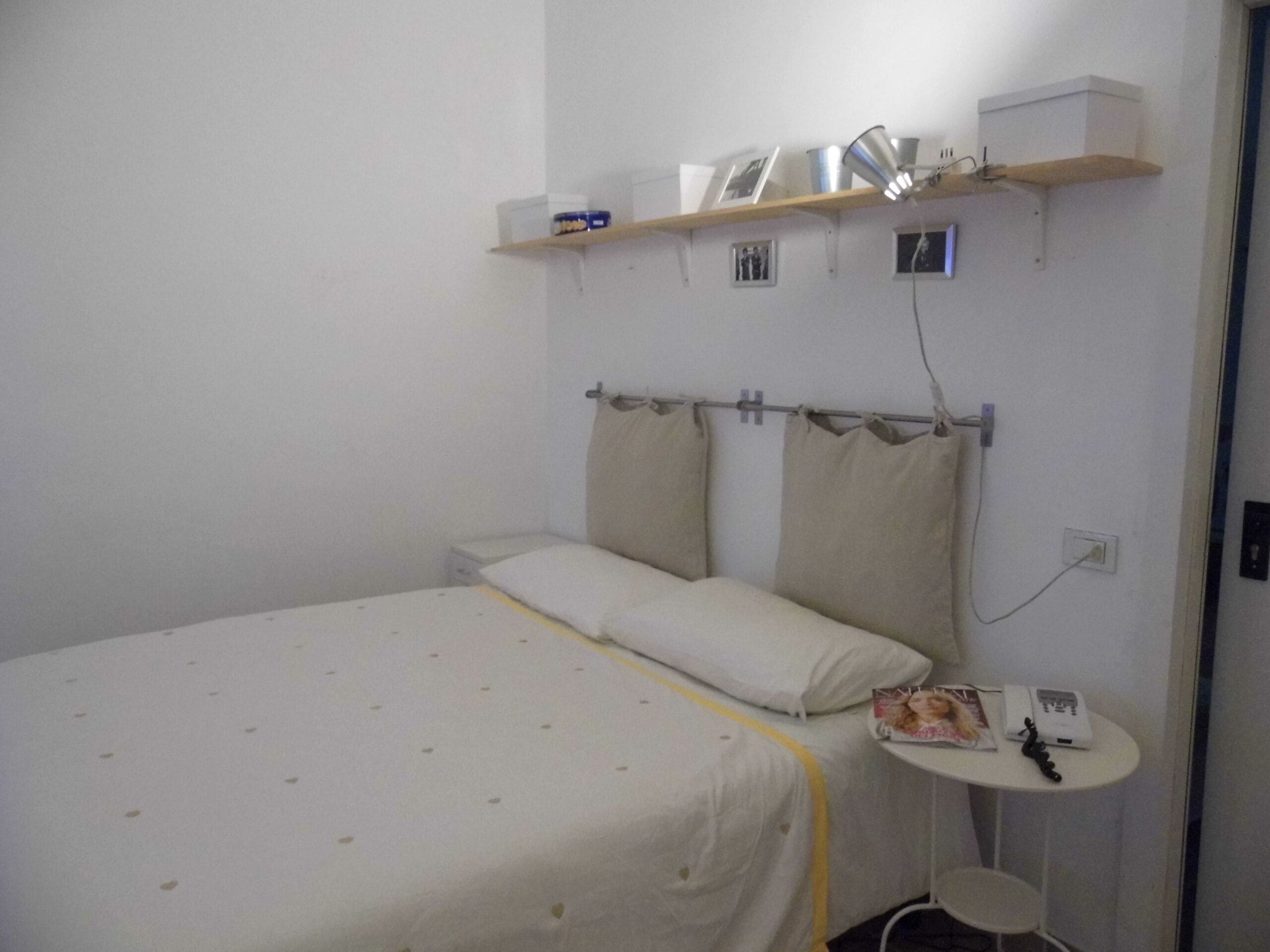Un letto singolo con copriletto bianco e giallo, un comodino con una lampada e un piccolo ripiano sopra il letto con alcuni oggetti.