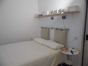 A single bed with a white and yellow bedspread, a bedside table with a lamp, and a small shelf above the bed with a few items.