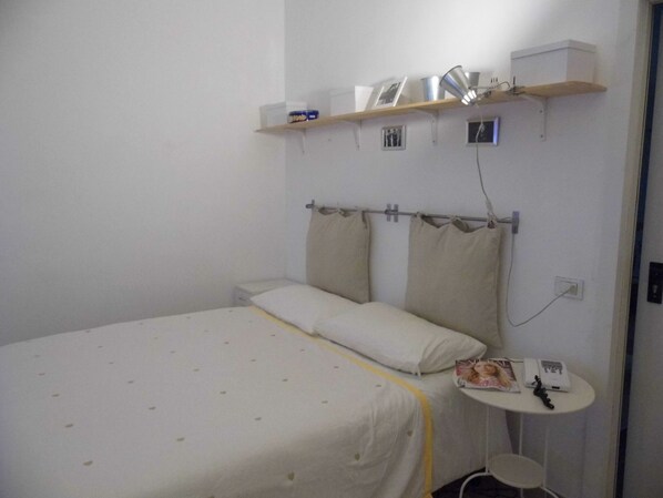 Una cama individual con una colcha blanca y amarilla, una mesita de noche con una lámpara y un pequeño estante sobre la cama con algunos objetos.