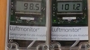 Zwei Geräte mit dem Aufdruck 'Luftmonitor', deren digitale Anzeigen Werte von 98,5 und 101,2 anzeigen.