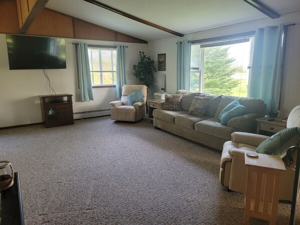 A living room with a beige sofa, a recliner, a small wooden side table, and a TV mounted on the wall. - Relaxing Lake View Home, Pet Friendly, Central Location. Close to Ferries! (St. Ignace)