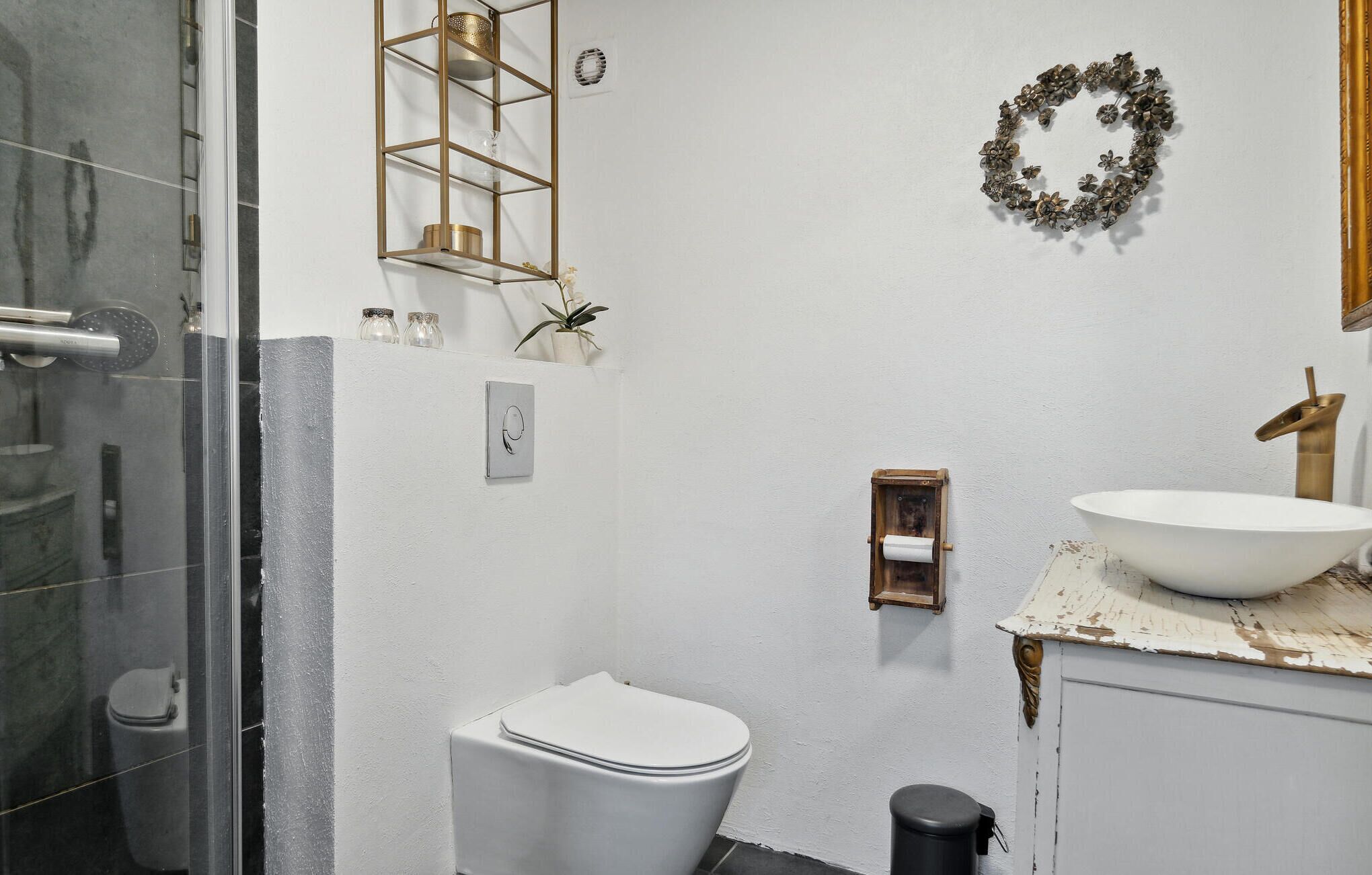 Ein Badezimmer mit Toilette in Weiß, einer Duschkabine aus Glas, einem weißen Waschbecken mit goldfarbenem Wasserhahn und einem Wandkranz.