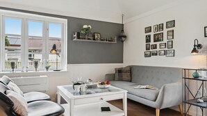 A modern living room with a grey sofa, a white coffee table, and a black leather chair. There are framed pictures on the wall and a window with a view of buildings outside. - 2 bedroom pet friendly home in ålbæk (Ålbæk)