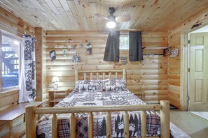 A log cabin bedroom with a bed, a nightstand, a lamp, a window with curtains, and wall decorations including fishing lures and a welcome sign. - Lakefront Phelps Cabin w/ Boat Dock & Water Toys! (Phelps)
