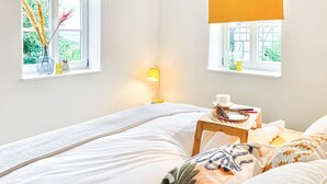 Une chambre à coucher avec un lit, une table de chevet avec une lampe, une fenêtre donnant sur un paysage verdoyant et un vase contenant des plantes séchées.