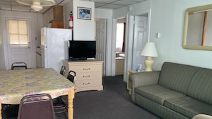 Quarto de hotel compacto com mesa de jantar, cadeiras, sofá, televisão, abajur e uma pequena cozinha.