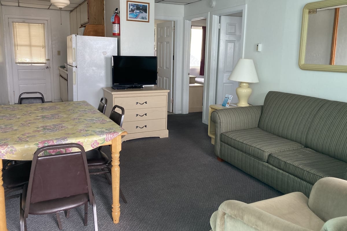 A compact hotel room with a dining table, chairs, a sofa, a television, a lamp, and a small kitchenette.