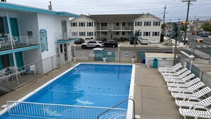 Piscina com design de tubarão, rodeada por cadeiras de descanso brancas e adjacente a um edifício com varanda.