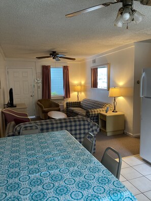 Une chambre d’hôtel de taille réduite, comprenant un lit, un canapé, une chaise, une petite table, un ventilateur de plafond et un réfrigérateur.