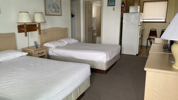 Quarto de hotel com duas camas, uma mesinha de cabeceira de madeira, uma escrivaninha, um frigorífico e uma janela com persianas.