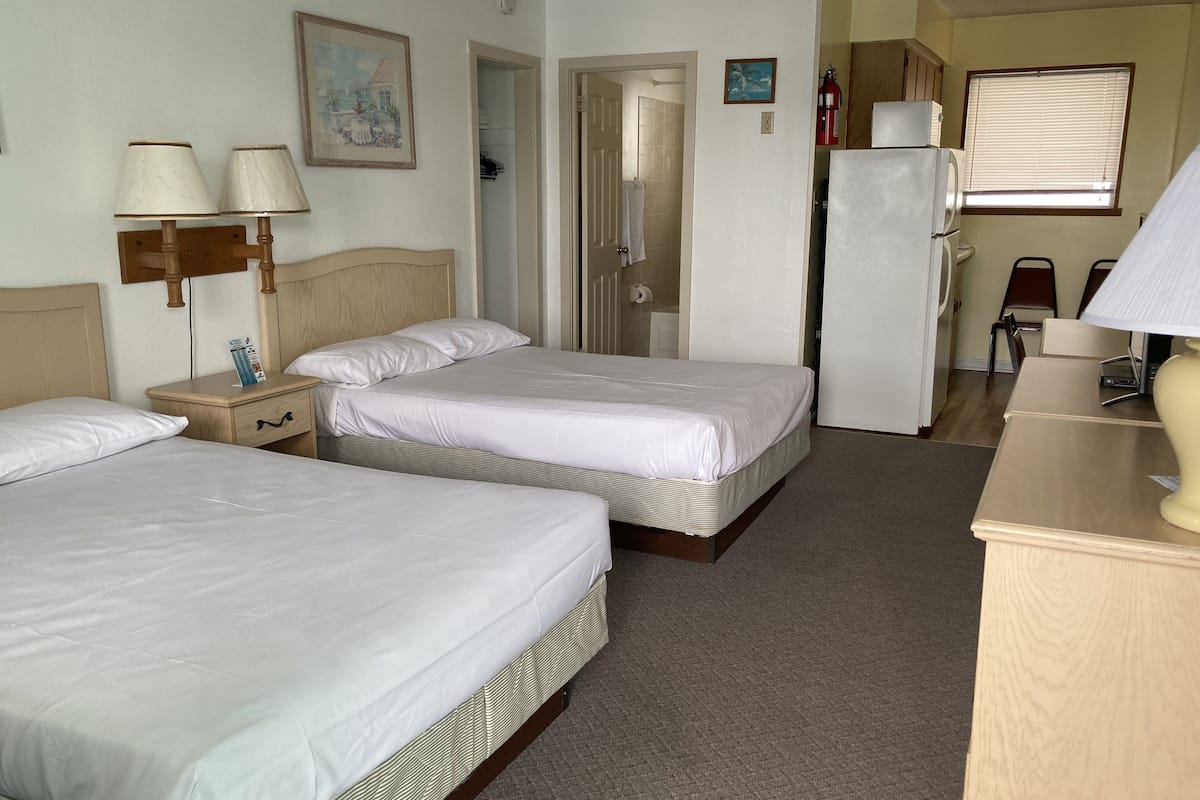 A hotel room with two beds, a wooden nightstand, a desk, a refrigerator, and a window with blinds.