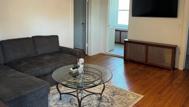 Un salon avec un canapé gris foncé, une table basse en verre agrémentée d’objets décoratifs, un plancher en bois, un ventilateur de plafond et un téléviseur à écran plat fixé au mur.