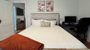 Une chambre à coucher comprenant un lit, un bureau avec un ordinateur, une chaise, un ventilateur de plafond et deux tableaux représentant des fleurs, accrochés au mur.
