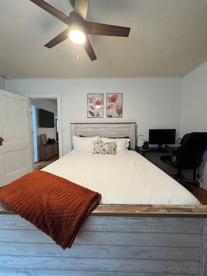 Une chambre à coucher comprenant un lit, un bureau avec un ordinateur, une chaise, un ventilateur de plafond et deux tableaux représentant des fleurs, accrochés au mur.