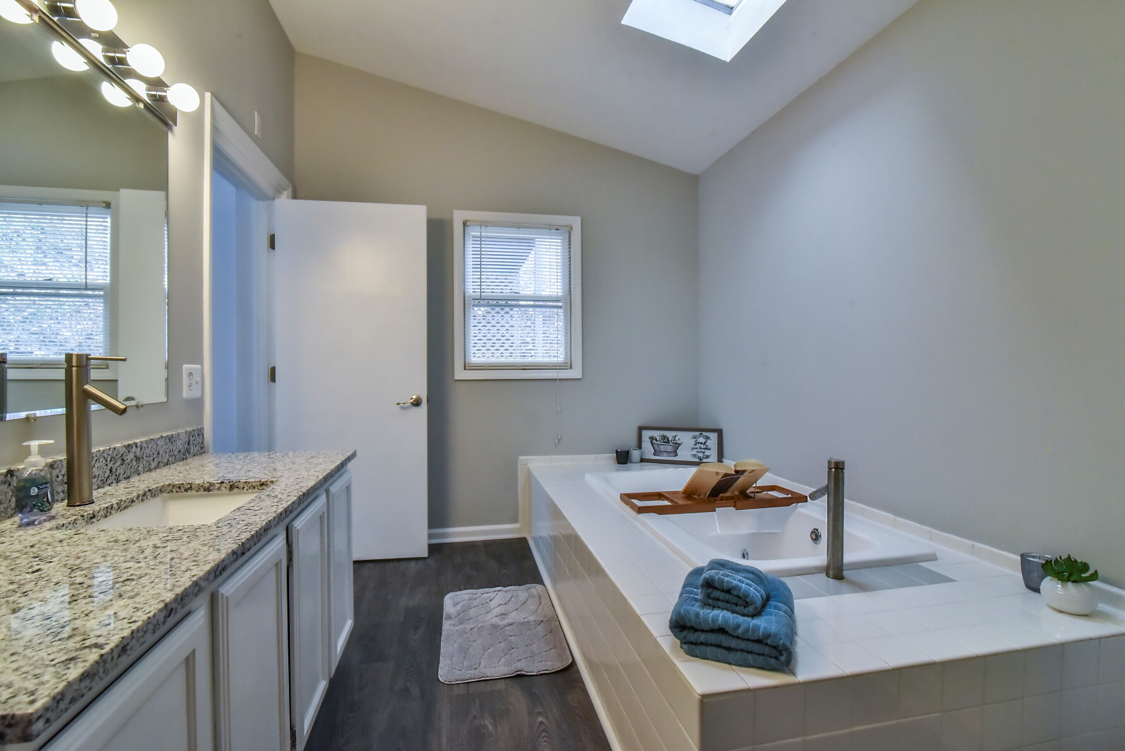 A modern bathroom with a large sink, a mirror with lights, a window with blinds, and a towel on the counter.