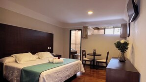Quarto de hotel com cama, uma mesinha com cadeiras, um vaso com flores e uma TV na parede.