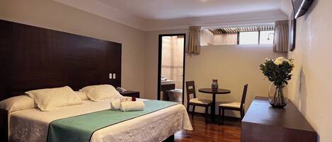 A hotel room with a bed, a small table with chairs, a vase with flowers, and a TV mounted on the wall.