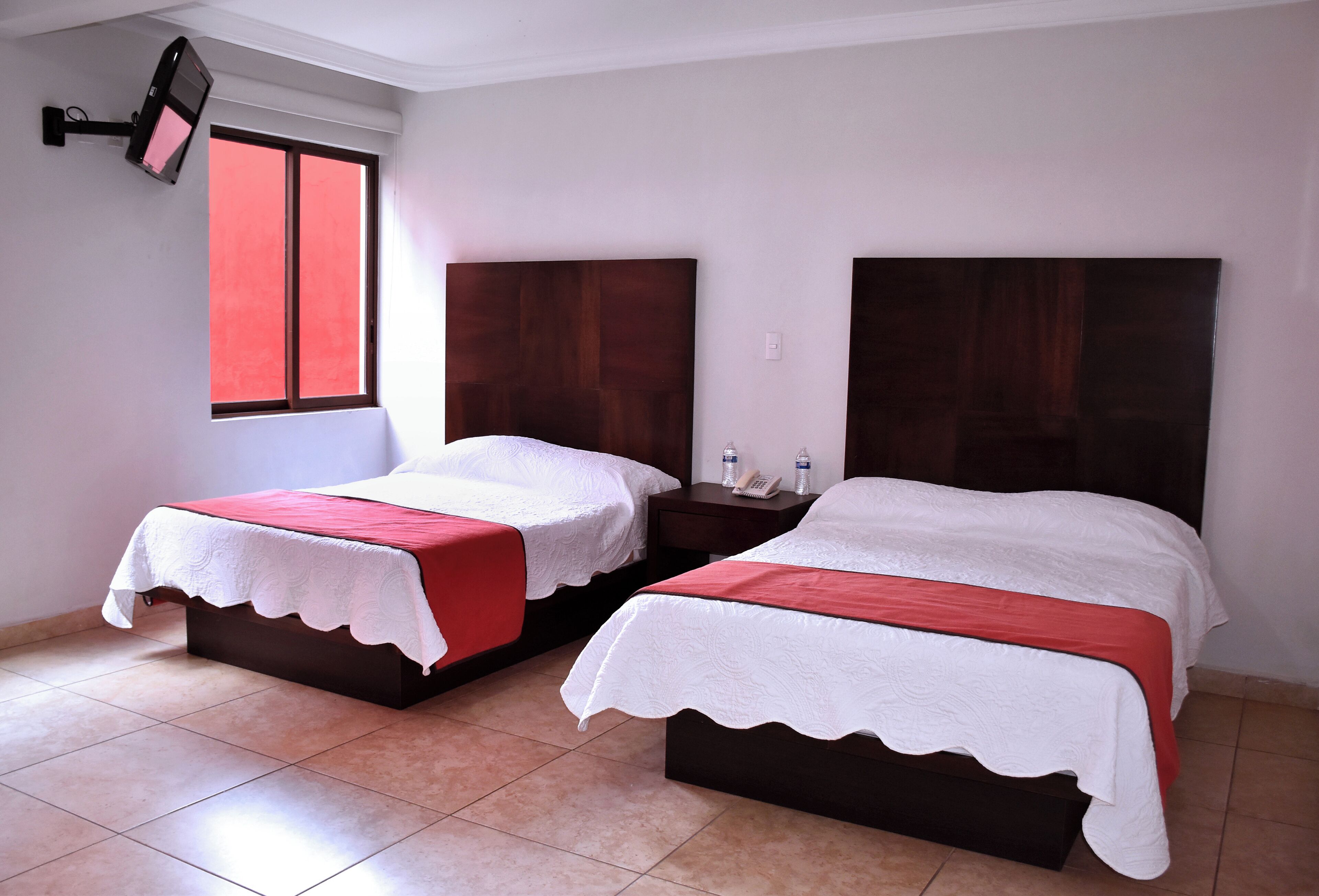Habitación de hotel con dos camas, una ventana con cortinas rojas, un televisor montado en la pared y una mesita de noche con un teléfono y una botella de agua.