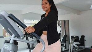 Uma pessoa usando uma bicicleta ergométrica numa academia, com diversos equipamentos de exercício ao fundo.