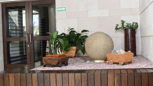 Uma área de recepção com um banco de madeira, plantas em vasos, uma esfera decorativa grande e um carrinho de madeira.