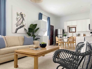 Una sala de estar con un sofá, una mesa de centro y sillas de mimbre. Se observa un comedor con una mesa y sillas. La cocina es visible, con armarios blancos y un refrigerador.