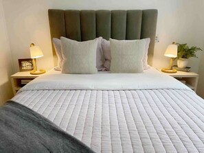 Cama bem arrumada com edredom branco acolchoado, dois travesseiros e cabeceira escura. Há duas mesas de cabeceira, com abajures e uma pequena planta em uma delas.