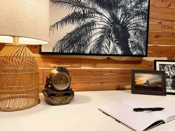 A wooden wall with a framed picture of palm leaves, a vintage-style clock, a wicker lamp, and a notebook with a pen on a white surface.