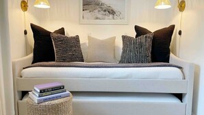 Un dormitorio moderno con un sofá blanco, dos lámparas de pared, un cuadro enmarcado sobre el sofá y una mesita de noche con libros.