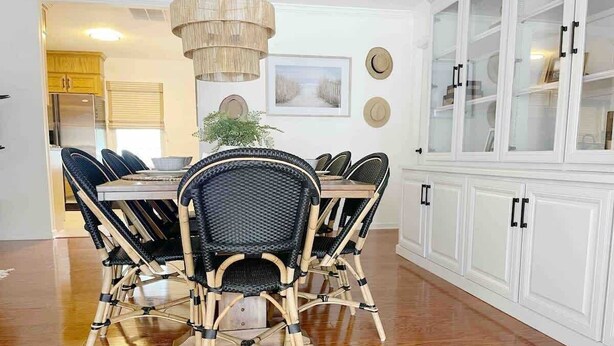 Un comedor con mesa de madera y sillas de mimbre, un armario blanco con puertas de cristal y una lámpara colgante.