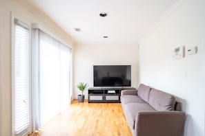 A modern living room with a flat-screen TV, a grey sofa, wooden flooring, and a potted plant.