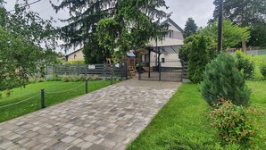 Ein gepflasterter Weg führt zu einem Gebäude mit überdachter Eingang, umgeben von Grünpflanzen und einem Zaun.