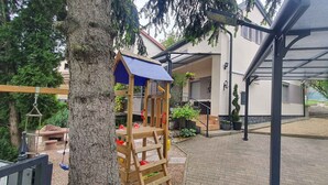 Ein überdachter Patio-Bereich mit einer Holz-Spielkonstruktion, einem überdachten Gang und einem Haus im Hintergrund.