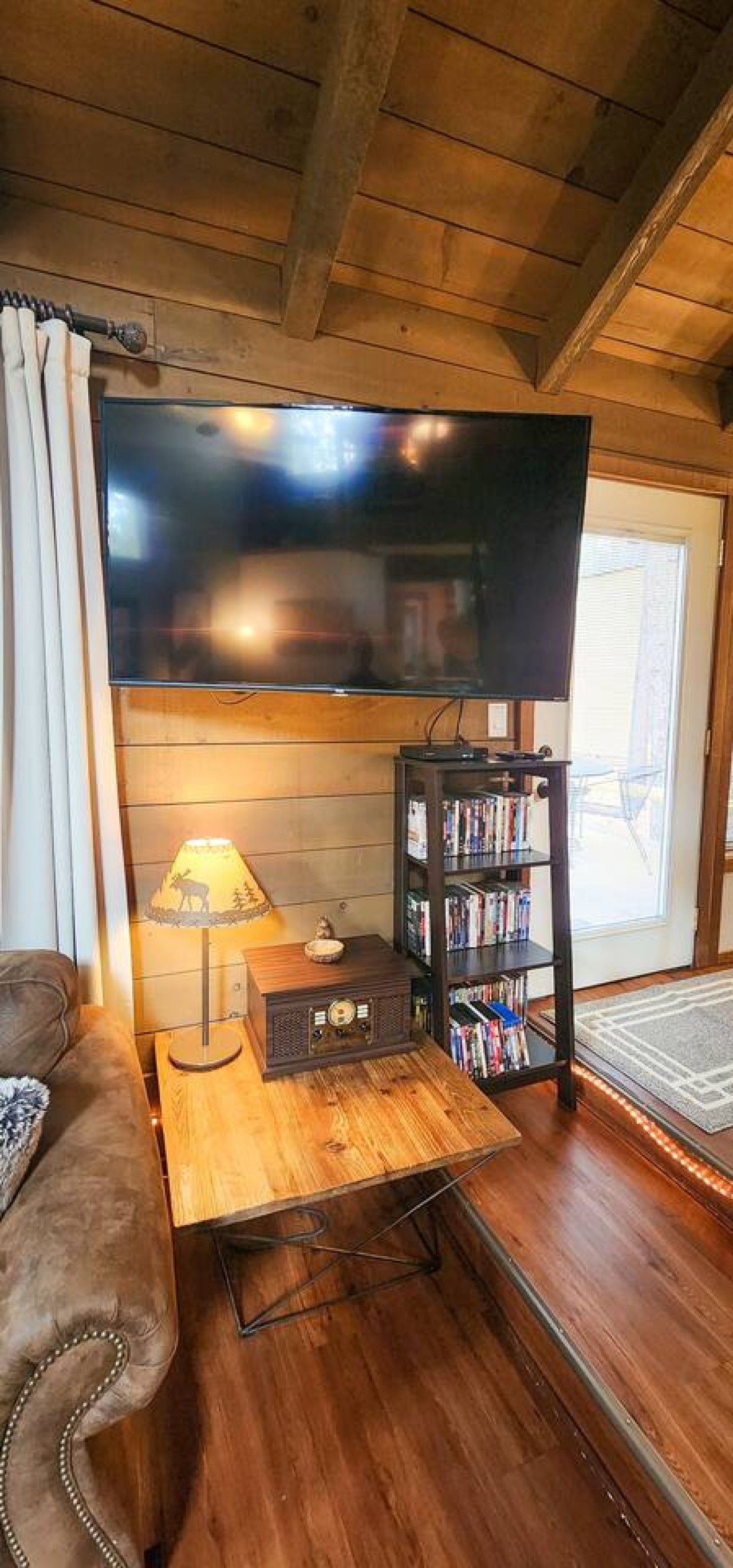 Une pièce chaleureuse avec un téléviseur à écran plat fixé au mur, une table basse en bois avec une lampe, une bibliothèque et un canapé confortable.