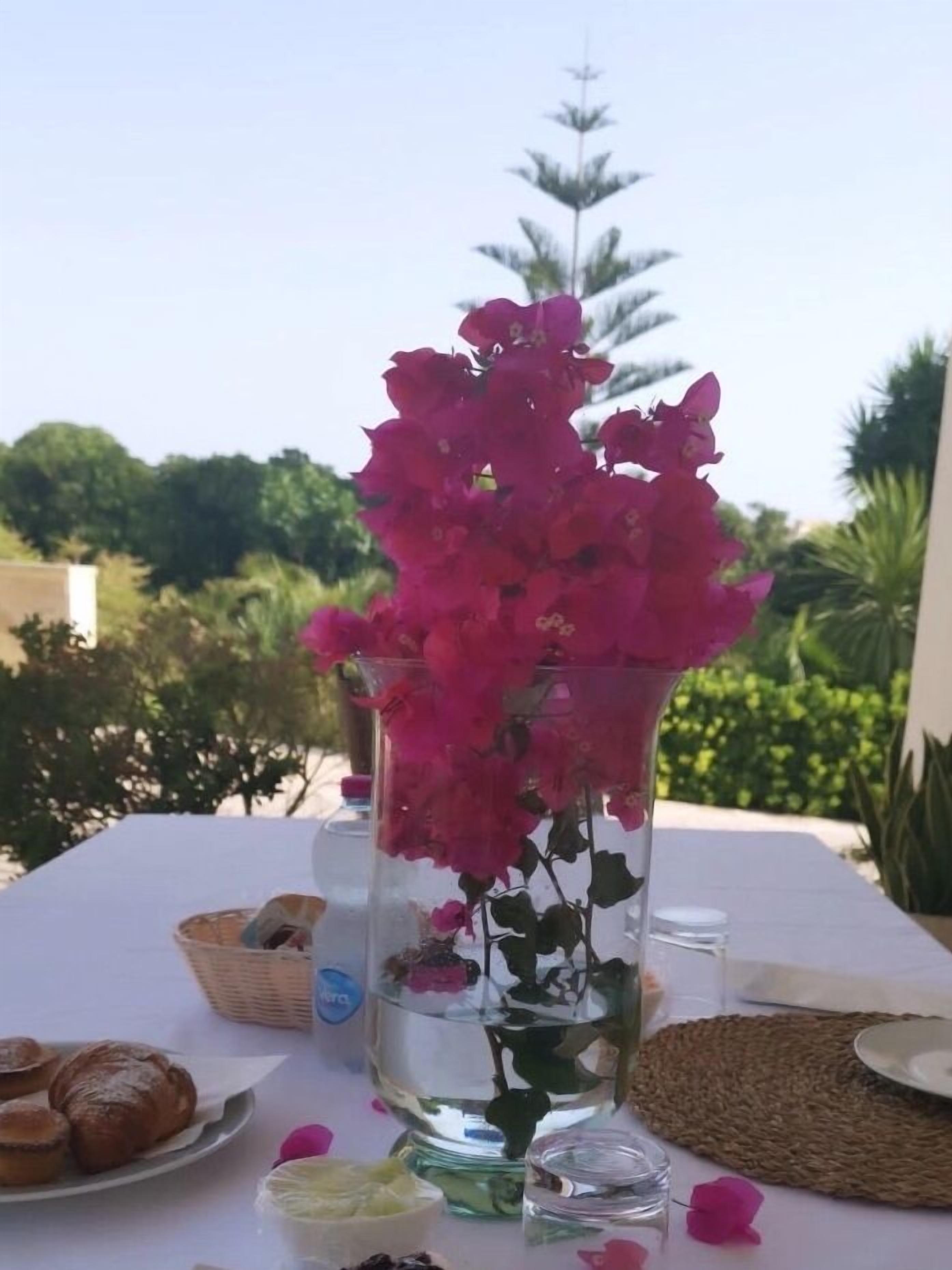 Ett bord med en vas med rosa blommor, en korg, en tallrik med bakverk och en liten skål med grädde.