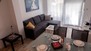 A modern living room with a black sofa, a glass dining table set for four, and a painting of sailboats on the wall.