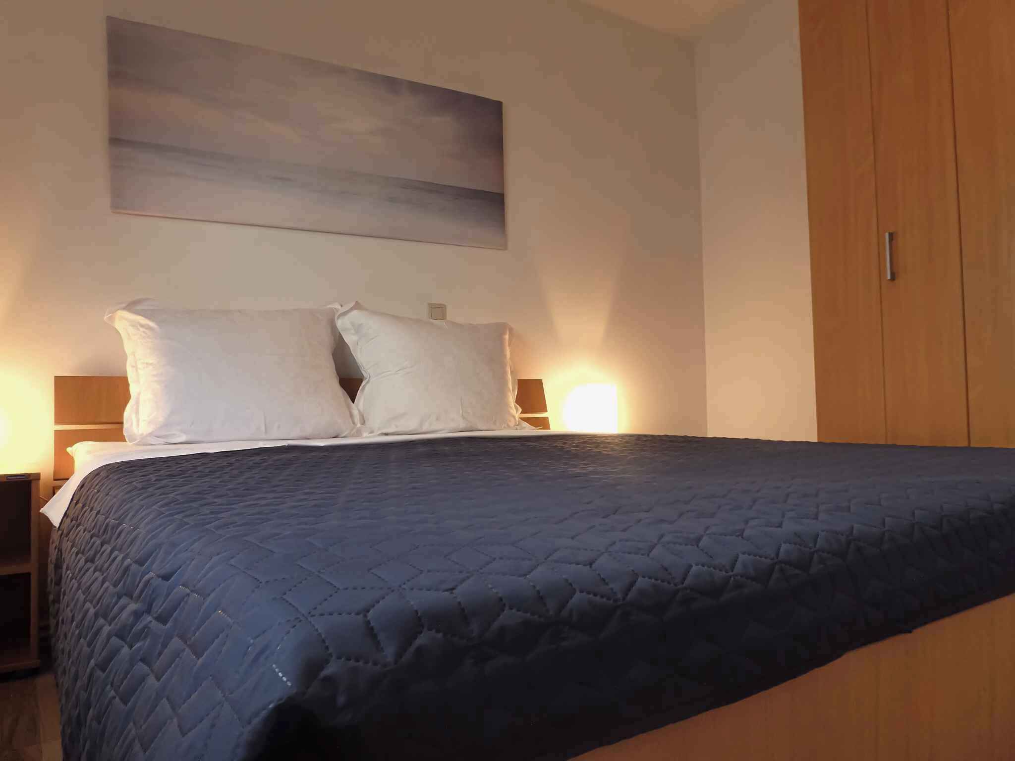 Un lit bien fait, recouvert d’une courtepointe sombre à motifs, avec deux oreillers, une armoire en bois sur le côté droit et un luminaire fixé au mur au-dessus du lit.