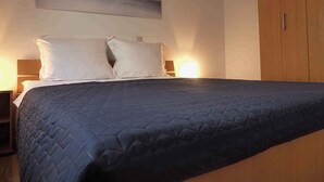A neatly made bed with a dark quilted bedspread and two pillows, a wooden wardrobe to the right, and a wall-mounted light fixture above the bed.