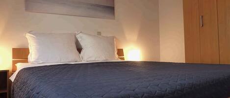 Un lit bien fait, recouvert d’une courtepointe sombre à motifs, avec deux oreillers, une armoire en bois sur le côté droit et un luminaire fixé au mur au-dessus du lit.