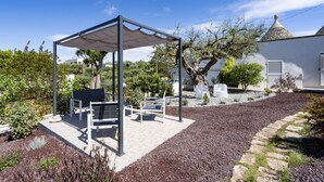 Ein überdachter Außenbereich mit Bänken, einer Pergola und einem Garten mit Kieswegen und Bäumen.