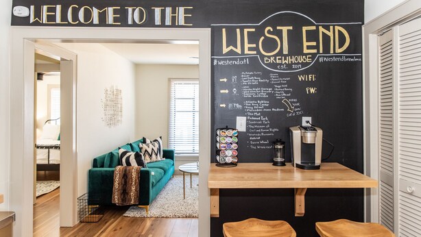 Un salon accueillant avec un canapé vert, une machine à café et un tableau noir affichant le menu du West End Brewhouse.