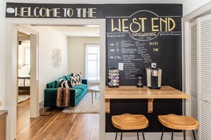 Un salon accueillant avec un canapé vert, une machine à café et un tableau noir affichant le menu du West End Brewhouse.